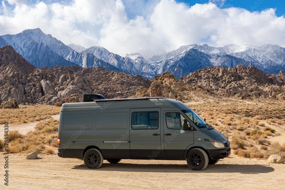 A dark gray camper van parked on a dirt road in a desert landscape with rocky hills and snow capped mountains in the background  Specialized RV Repair and Conversions, Inc 