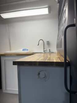 sprinter-conversions_0000_IMG_0704 A kitchen workspace with a wood countertop, stainless steel faucet, sink, and a fluorescent light fixture on the wall above. Specialized RV Repair and Conversions, Inc.