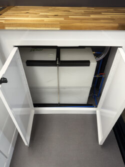 sprinter-conversions_0002_IMG_0702 A large white water tank with connected pipes is installed in a cabinet under a wooden countertop with the cabinet doors open. Specialized RV Repair and Conversions, Inc.