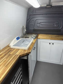 sprinter-conversions_0003_IMG_0701 Interior of a mobile workspace with wooden countertops, white cabinets, a sink, and packaged items on a scale under bright overhead lighting. Specialized RV Repair and Conversions, Inc.