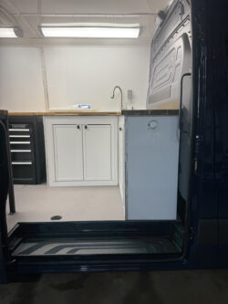 sprinter-conversions_0005_IMG_0696 View of a van conversion interior with a countertop, sink, white cabinets, drawer unit, and overhead lighting, seen from the open side door. Specialized RV Repair and Conversions, Inc.