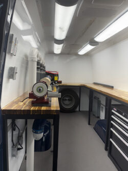 sprinter-conversions_0007_IMG_0709 Interior of a mobile workshop with wooden countertops, a mounted grinder, tool drawers, storage shelves, and a spare tire against the back wall. Specialized RV Repair and Conversions, Inc.